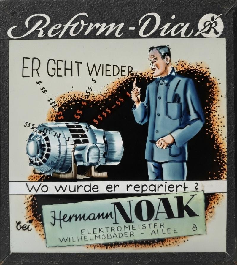 Farbiges Kinodia: Ein Mann zeigt auf einen Elektromotor, darunter „Wo wurde er repariert? Bei Hermann Noak, Elektromeister, Wilhelmsbader-Allee 8“